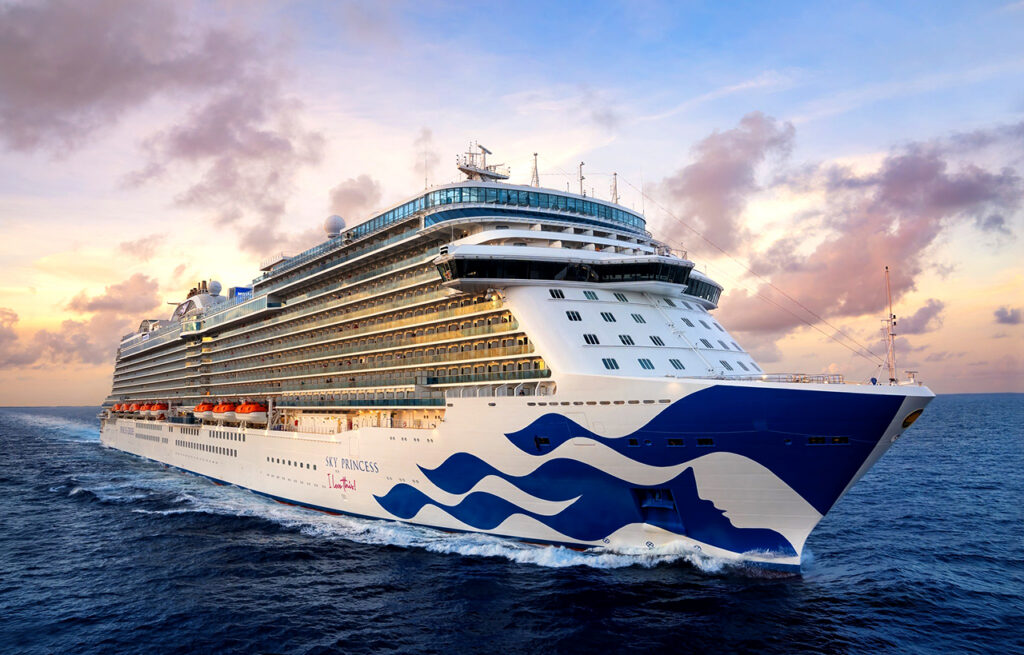 An exterior side-view of the Sky Princess cruise ship sailing across the ocean during a golden sunset, featuring its iconic blue wave hull art. This vessel offers a traditional luxury experience that rivals a celebrity cruise, with expansive balconies and premium amenities for a sophisticated voyage.