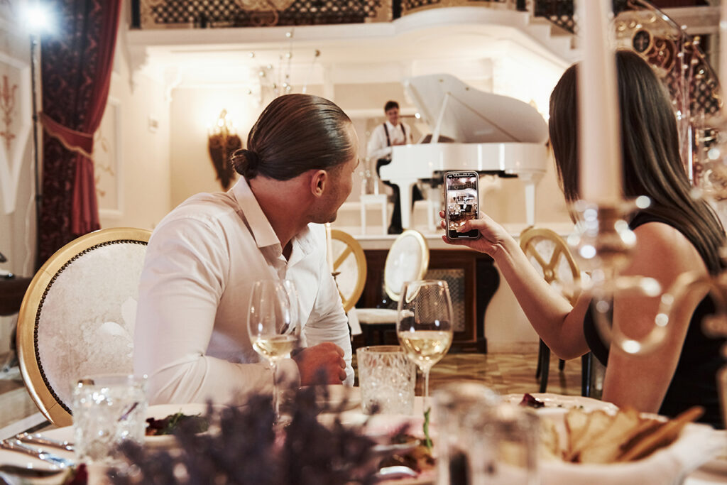 A couple dines at a candlelit table in an elegant cruise ship restaurant, attentively listening to a pianist playing a grand piano in the background. This sophisticated evening atmosphere is a central point in the princess vs royal caribbean comparison, as travelers evaluate cruise quality and refined entertainment options when considering overall cruise pricing for their next vacation.