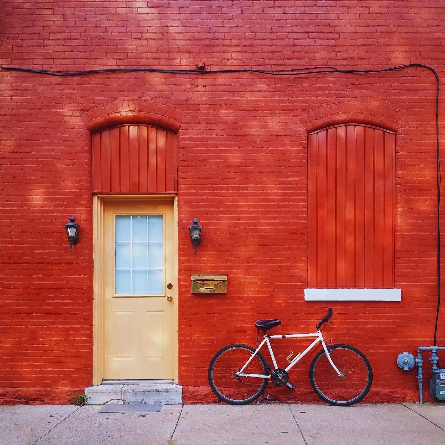 A white bicycle parked against a vibrant red brick wall next to a pale yellow door and shuttered window, a charming urban scene for those applying experience to capture minimalist street photography.