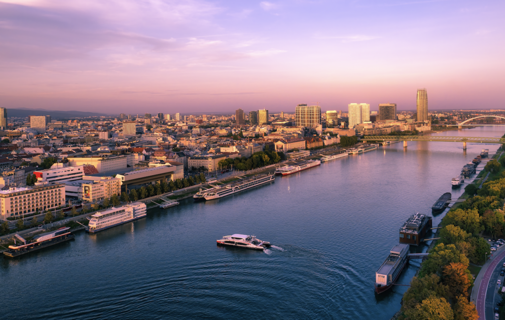 A sleek river cruise ship sailing past a dramatic, modern city skyline at dusk, with towering glass skyscrapers and their bright lights reflecting on the dark river water.