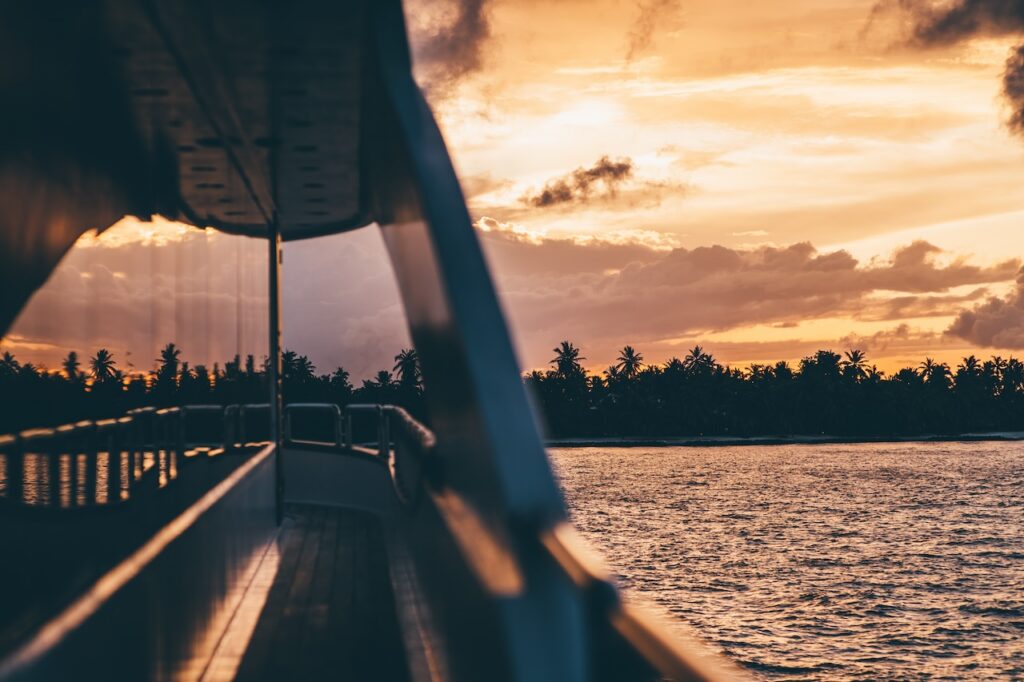 Golden hour sunset viewed from the deck railing of a moving yacht or boat, showing silhouetted palm trees on a tropical island shoreline and reflecting light on the ocean water.