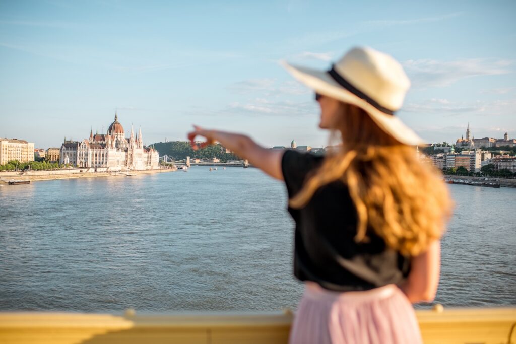 Solo traveler in a sun hat pointing toward the iconic Hungarian Parliament Building in Budapest, representing independent exploration on a river cruise.