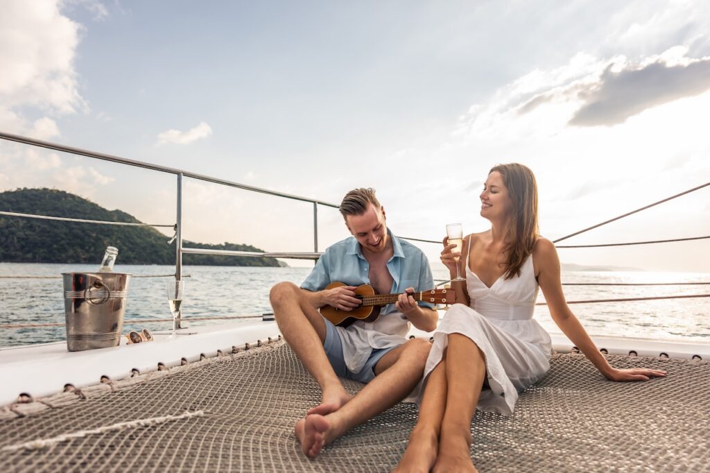 "A smiling, barefoot couple lounges on a catamaran's netting over the ocean at sunset; the man plays a ukulele while the woman holds a glass of champagne, depicting a relaxed, romantic cruise.
