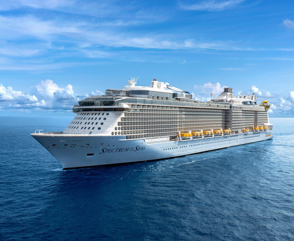 A grand wide-angle view of the Allure of the Seas sailing in open water, perfectly illustrating the architectural scale of the Oasis Class—one of the most famous Royal Caribbean ship classes. The image captures the ship's split-hull design and the iconic AquaTheater at the aft, representing the high-energy and innovative features that define this specific class of vessels.