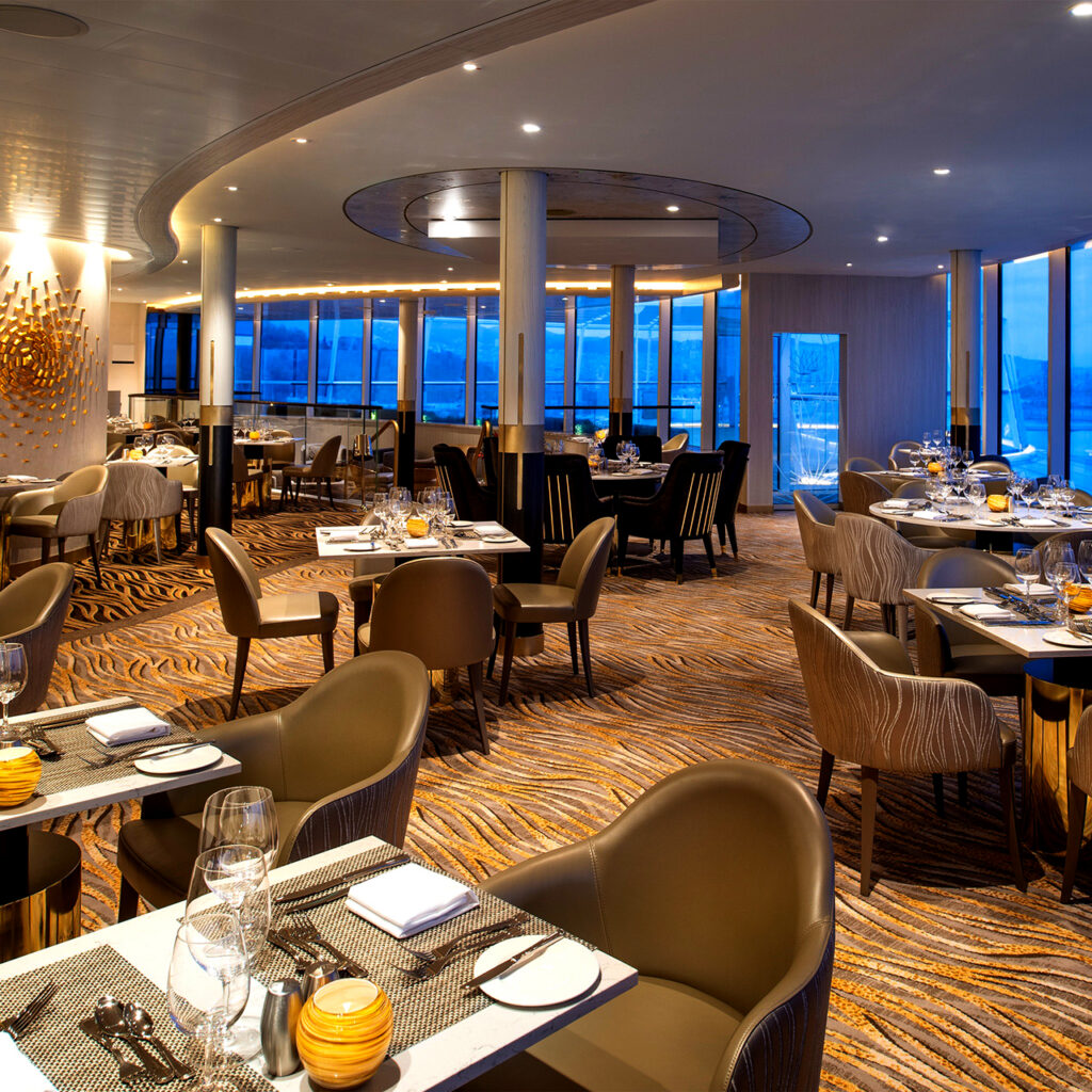 A sophisticated specialty restaurant onboard a luxury ship featuring floor-to-ceiling windows, modern gold-accented pillars, and plush seating. The refined interior design and panoramic ocean views represent the exclusive amenities found within the Royal Caribbean suites neighborhood.