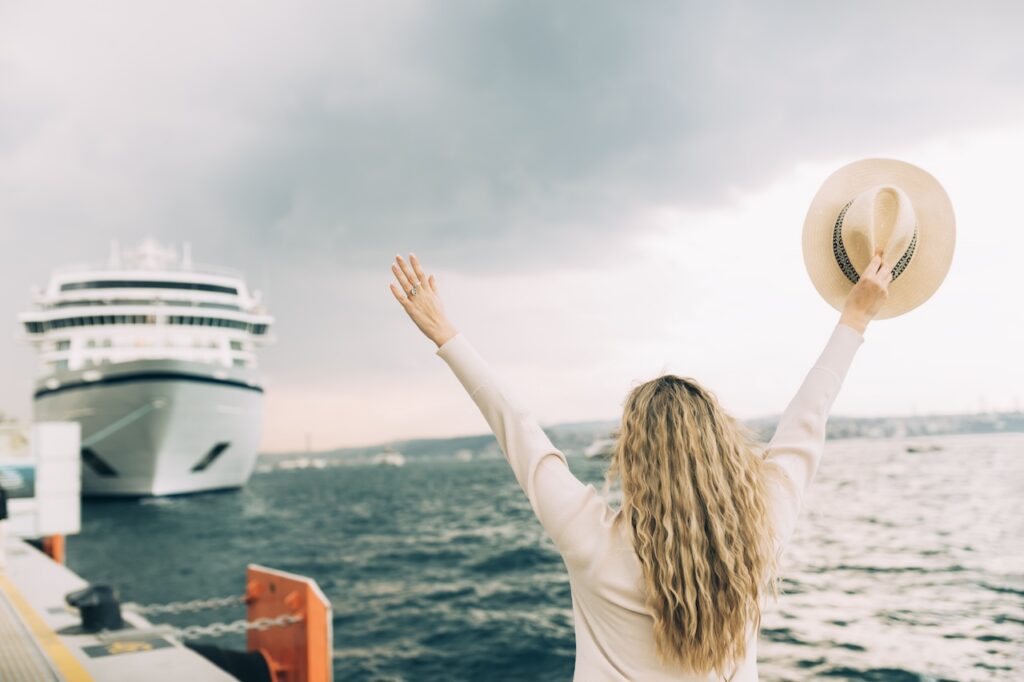 A woman with long blonde hair, seen from behind, stands at a port dock with her arms raised in triumph or farewell, holding a straw hat, as a large white cruise ship is visible in the background over the deep blue water.