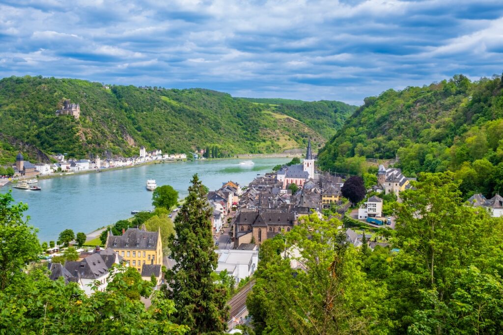 High-angle aerial view of a picturesque German river town nestled in a valley, with the Rhine River winding through steep, lush green, forested hills and a prominent church steeple rising from the red-roofed buildings.
