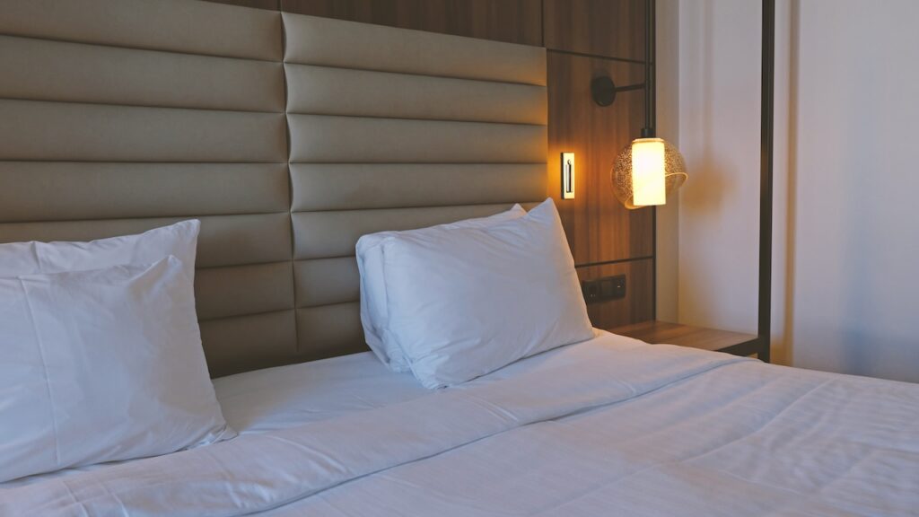 Close-up of a modern, well-made bed in a luxurious hotel room or a scenic cruise line cabin, featuring clean white linens, plump pillows, a padded tan headboard, and warm ambient lighting from a wall sconce.