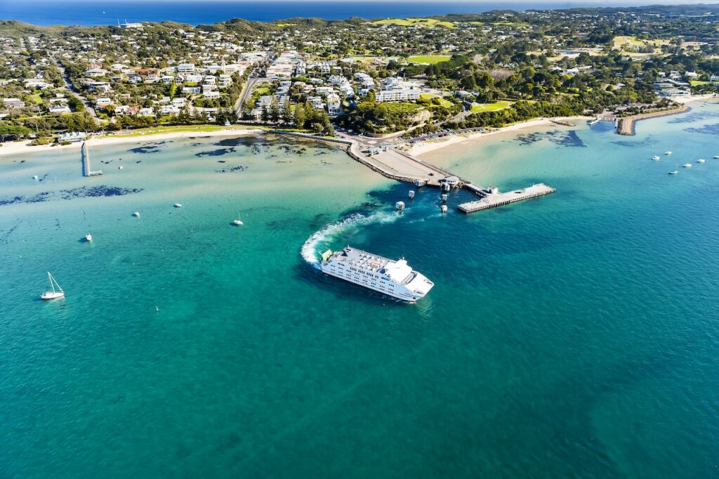 Aerial view of a white passenger ferry or small cruise ship turning in bright turquoise water near a pier and the coastline, characteristic of a scenic cruise line Australia port or departure.