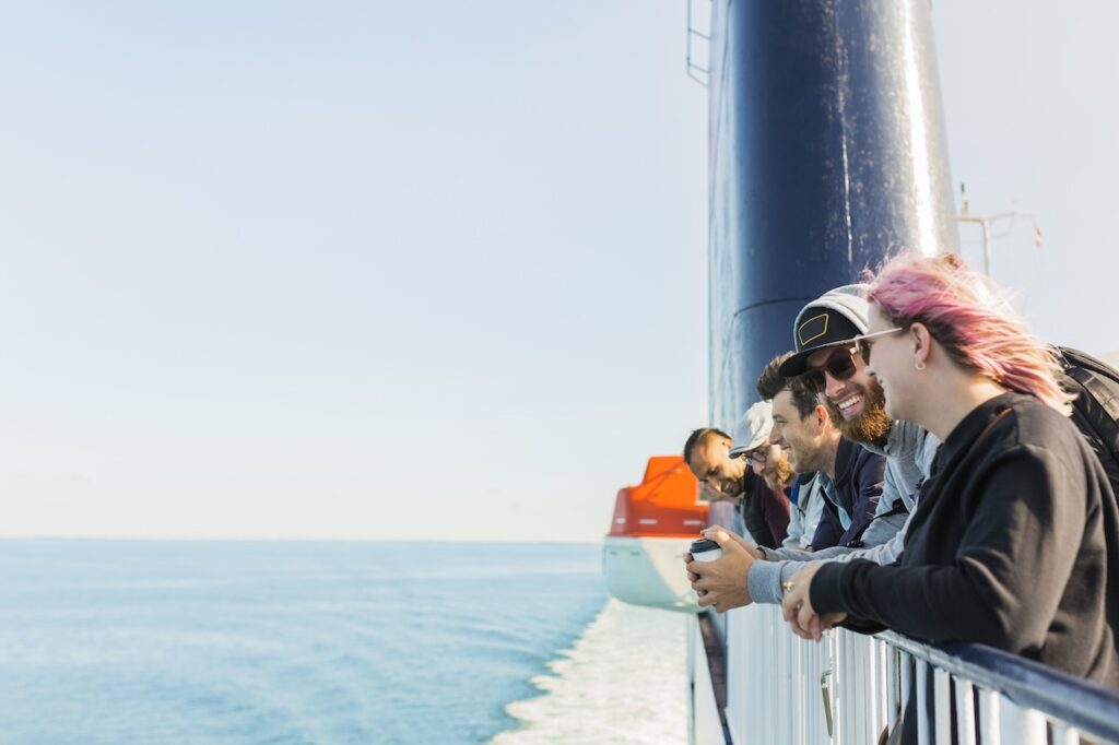 A diverse group of friends smiling and leaning against the ship's railing, looking out over the vast, calm blue ocean on a sunny day. One person has pink hair and another holds a coffee cup.