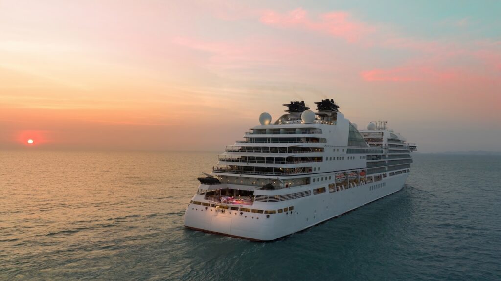 An aerial or low-angle view of a large, modern white cruise ship sailing on the ocean during a vibrant sunset, with the sun low on the horizon casting warm orange and pink hues across the sky and water.