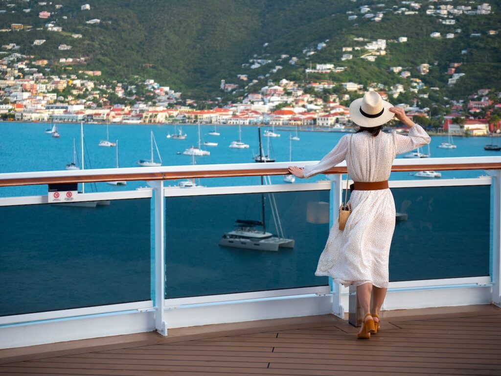 A woman wearing a white sun dress and a straw hat stands on a ship deck or balcony, looking out over a busy harbor filled with sailboats and a colorful, terraced coastal city rising in the background.