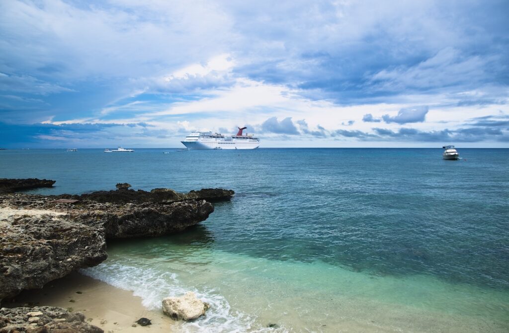 A large white cruise ship anchored in clear, turquoise water near a rocky shoreline and sandy beach under a dramatic blue sky, representing an exotic port stop on a scenic ocean cruise line itinerary.