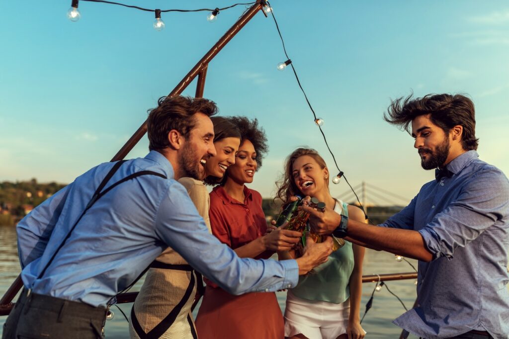 A diverse group of five smiling friends cheering and clinking bottled drinks together on the deck of a boat at sunset, celebrating and enjoying a lively social outing or party.