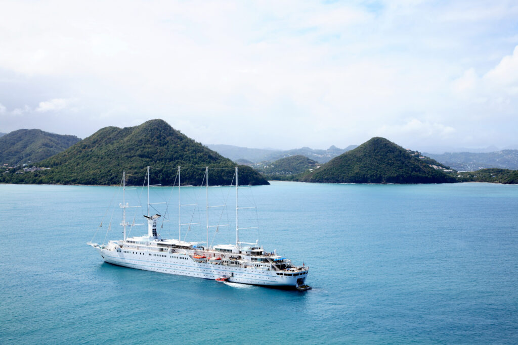 A large, elegant white sailing cruise ship with four tall masts anchored in a bright turquoise bay, representing a popular cruise ship port in Tortola, British Virgin Islands. Lush, green tropical mountains rise from the coastline under a soft, cloudy sky.