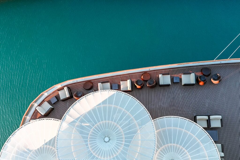 High-angle aerial view looking straight down onto the wooden deck of a cruise ship, showing a curved arrangement of lounge chairs, small tables, and sun shades beside the bright turquoise water of the ocean or a port.