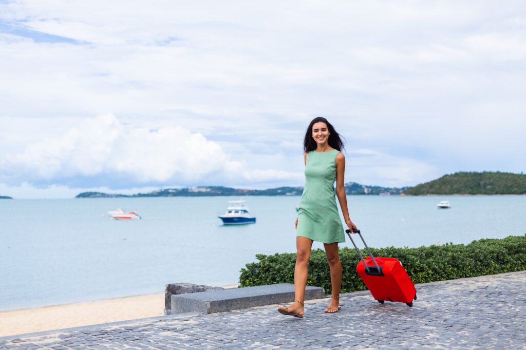 A solo traveler on a sunny beach promenade with a single piece of luggage, looking relaxed and ready to explore her destination.