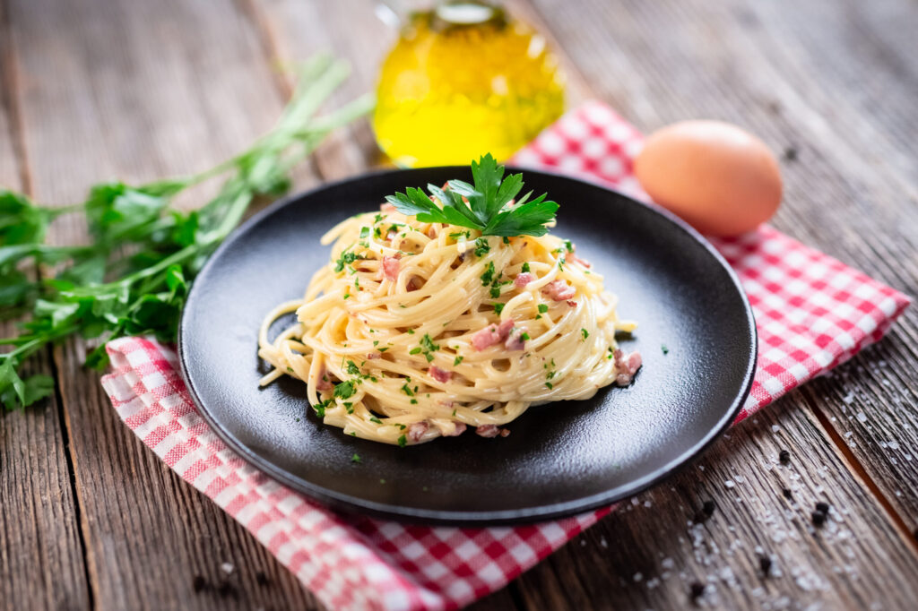 A plated serving of creamy Spaghetti alla Carbonara topped with pancetta and fresh parsley on a black plate, set against a rustic wooden table with olive oil and an egg.