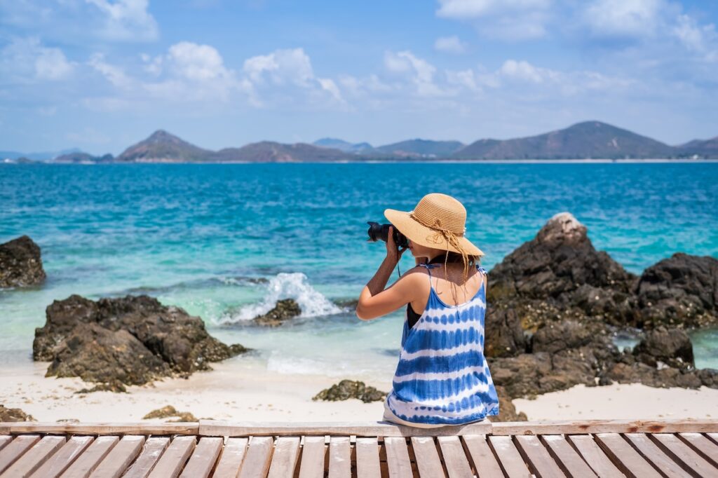 Woman in a sun hat taking a scenic photo of the turquoise ocean and mountainous islands on a rocky tropical beach, highlighting travel photography and beautiful views.