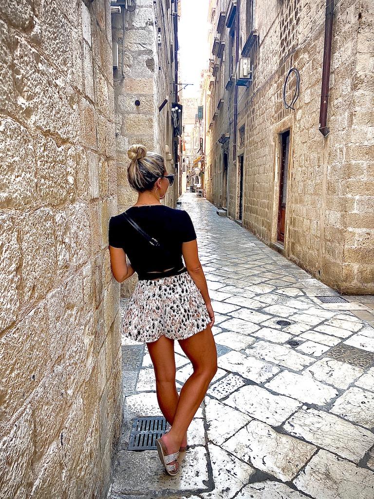A woman in a black shirt and patterned shorts stands in a narrow cobblestone alleyway, capturing the historic atmosphere of old town Dubrovnik.
