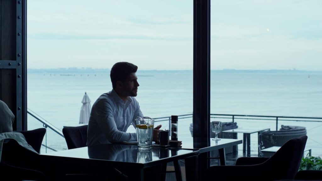 A man in a white dress shirt sits at a table in a luxurious hotel restaurant, looking out of large floor-to-ceiling windows toward a calm ocean view.