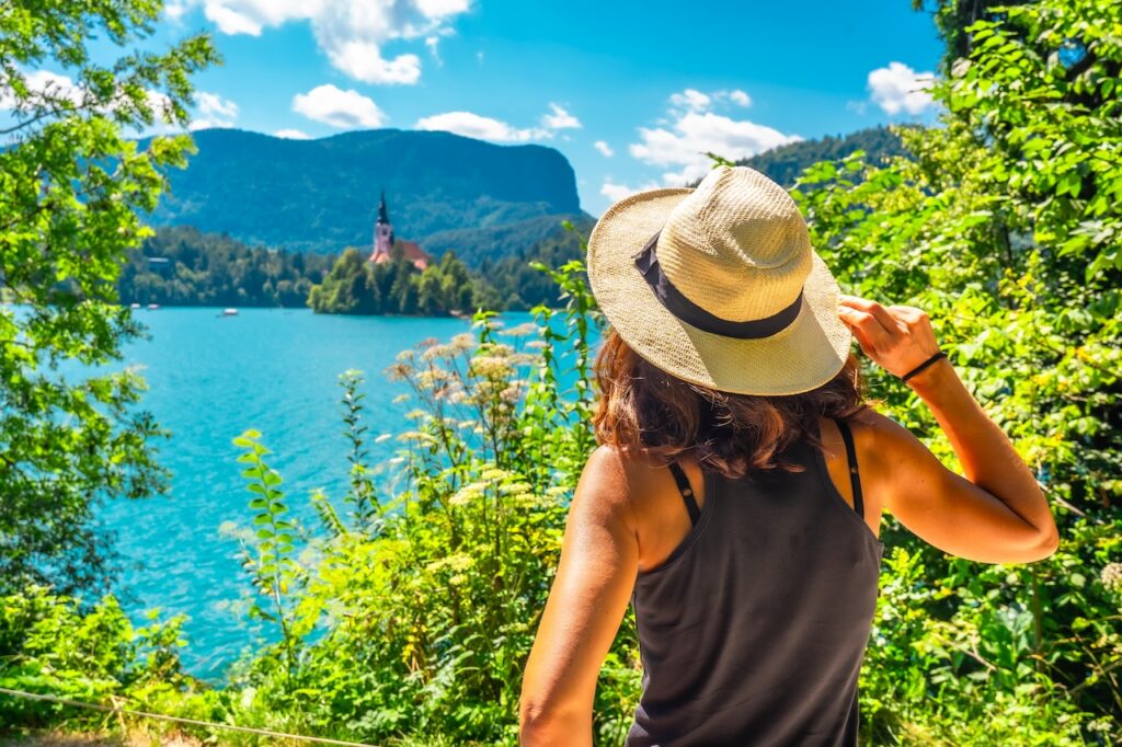 A traveler wearing a sunhat looks out over the bright blue water and island church at Lake Bled, surrounded by lush green trees on a sunny day.