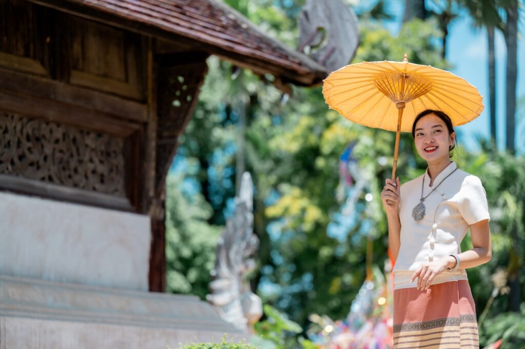 Woman in traditional Thai clothing holding a yellow umbrella at a temple in Thailand.