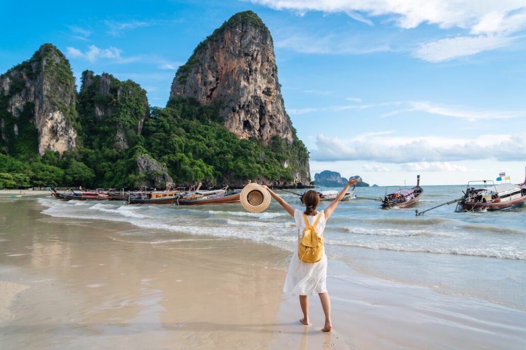 Traveler standing on a stunning beach with traditional longtail boats and iconic limestone cliffs, highlighting a top must-see destination in Thailand.