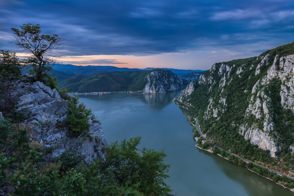 Dramatic twilight view of the Danube River flowing through the steep, rocky cliffs of the Iron Gates Gorge, with a winding road visible on the mountain base.