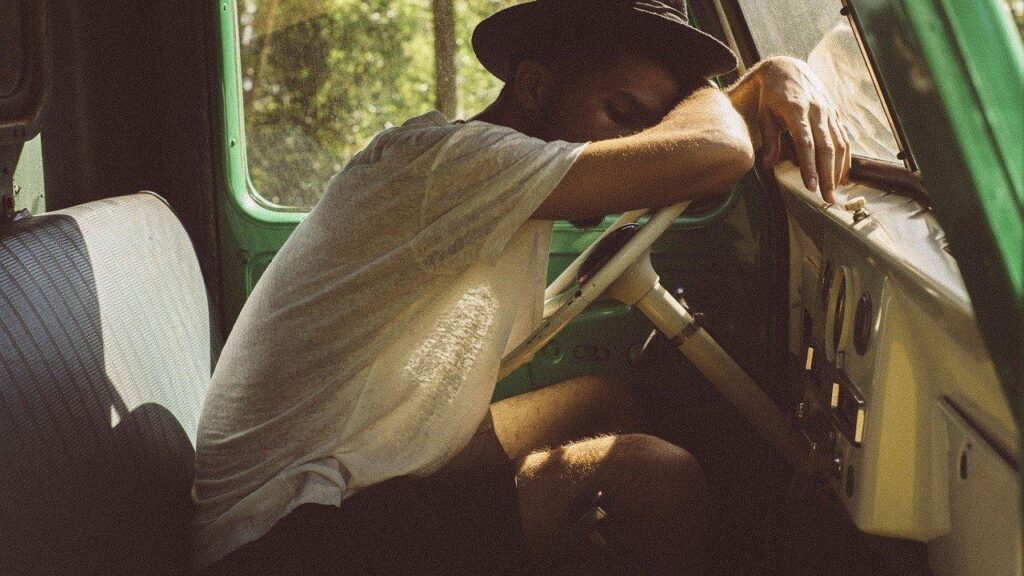 A man in a fedora resting his head on the steering wheel of a vintage green truck, capturing a candid moment of road trip fatigue and the importance of travel tips for long journeys.