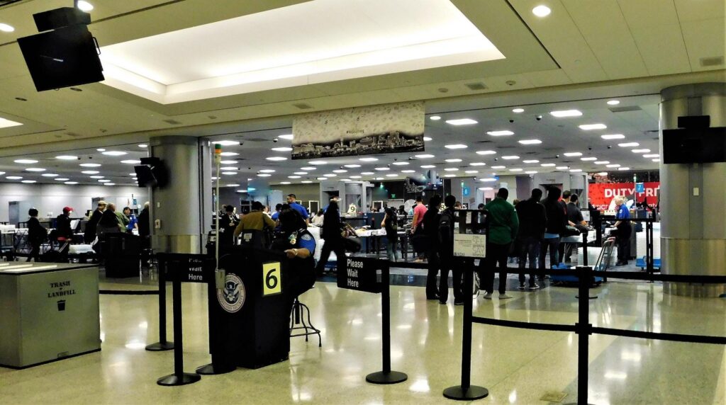 Passengers queue at an airport security checkpoint with TSA officers and screening equipment in a bright terminal.