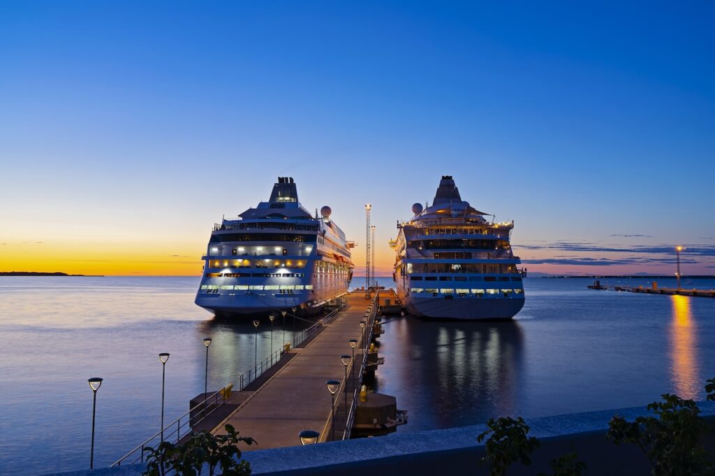 Two large illuminated cruise ships docked at a pier during a colorful sunset, highlighting a premium travel experience and evening port arrival.