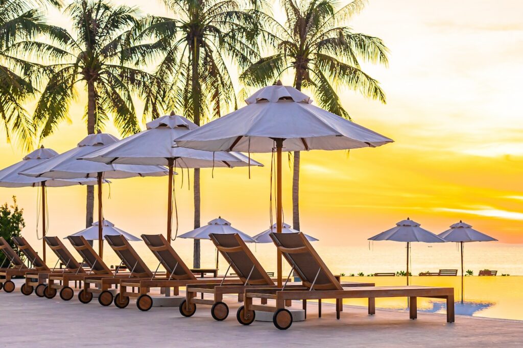 Luxurious beach resort view featuring thatched beach huts, a turquoise pool, and tropical palm trees, highlighting a private and relaxing getaway.