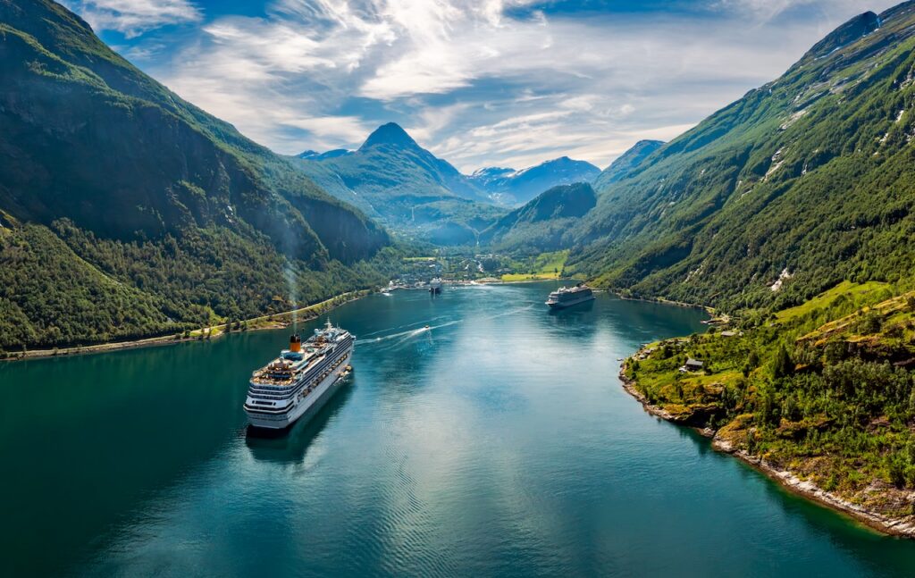 Aerial view of a large white and orange cruise ship navigating a narrow, calm, deep-water fjord. The ship is dwarfed by the dramatic, heavily forested, sheer mountain slopes rising sharply on both sides under a bright, sunny sky.