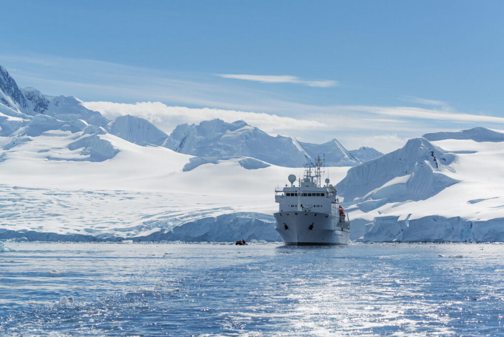 One of the modern cruise ships sailing through the icy waters of Antarctica, surrounded by massive snow-covered mountains and glaciers under a bright blue sky.