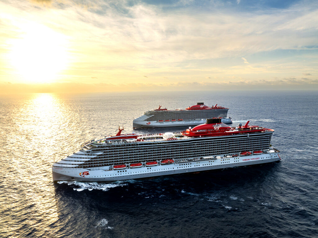 An aerial side-view of two sleek Virgin Voyages cruise ships sailing side-by-side on the open ocean at sunset. The modern gray hulls and iconic red funnels stand out against the shimmering golden water, showcasing the upscale atmosphere of an adults-only cruise.