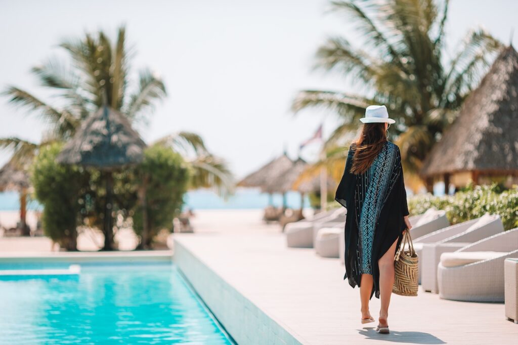 A woman wearing a wide-brimmed hat, a dark cover-up, and carrying a woven bag walks along the edge of a turquoise swimming pool at a sunny tropical beach resort. Palm trees and thatched-roof structures are visible in the background.
