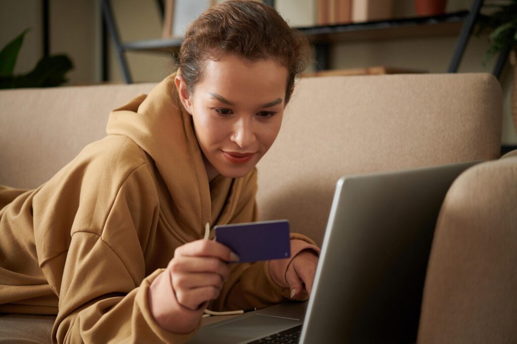 A woman in a tan hoodie lies comfortably on a sofa, using a laptop and holding a credit card to book a trip or manage rewards through the capital one travel portal.
