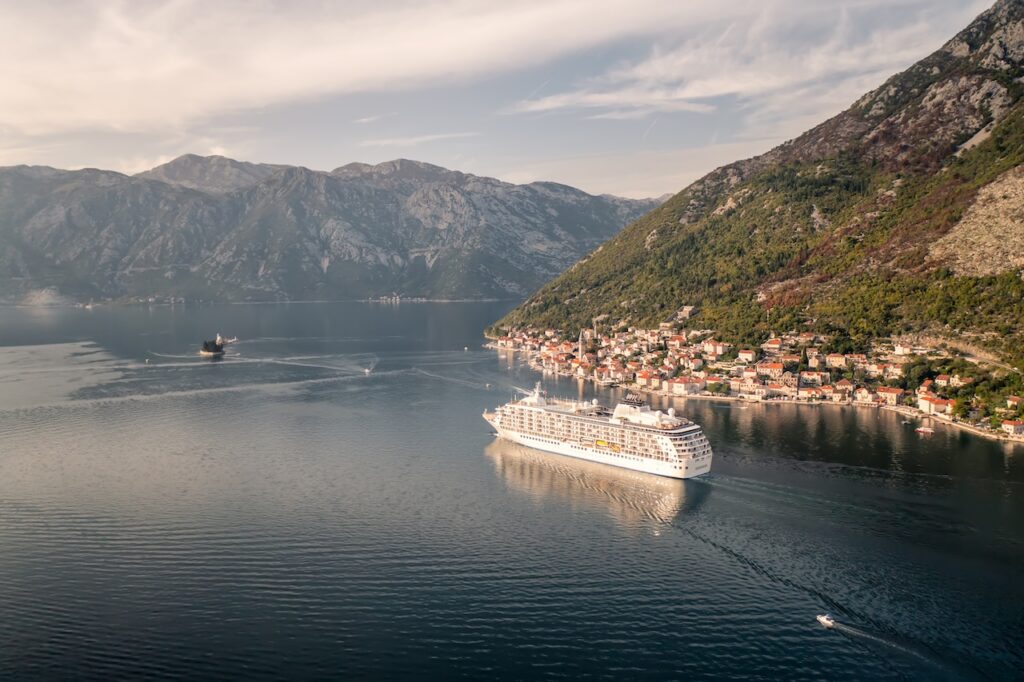 Luxury cruise ship sailing past a historic coastal town and steep mountains, representing a unique destination stop on the Silversea 2027 World Cruise.